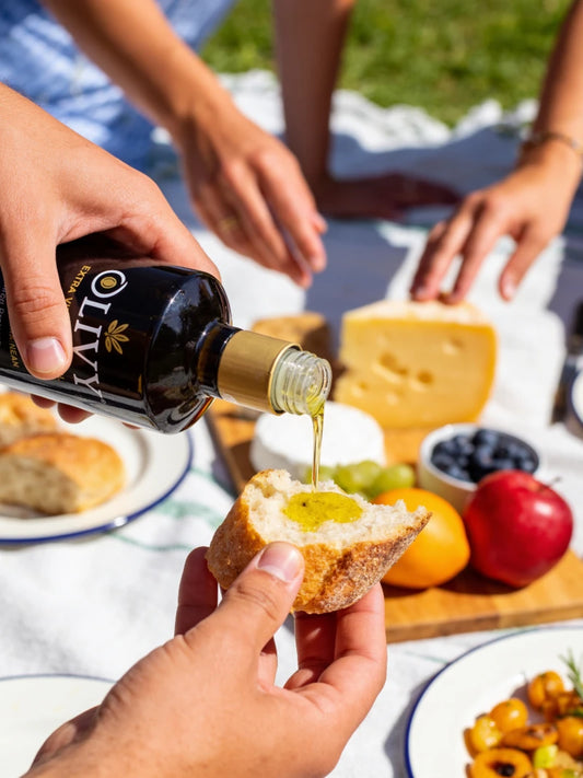 A close-up lifestyle shot of a picnic scene where a hand holds a slice of bread while another pours olive oil from a bottle labeled 'OLIVY' onto it. A wooden board with cheese, blueberries, an orange, and a red apple sits on a white picnic blanket in the background.