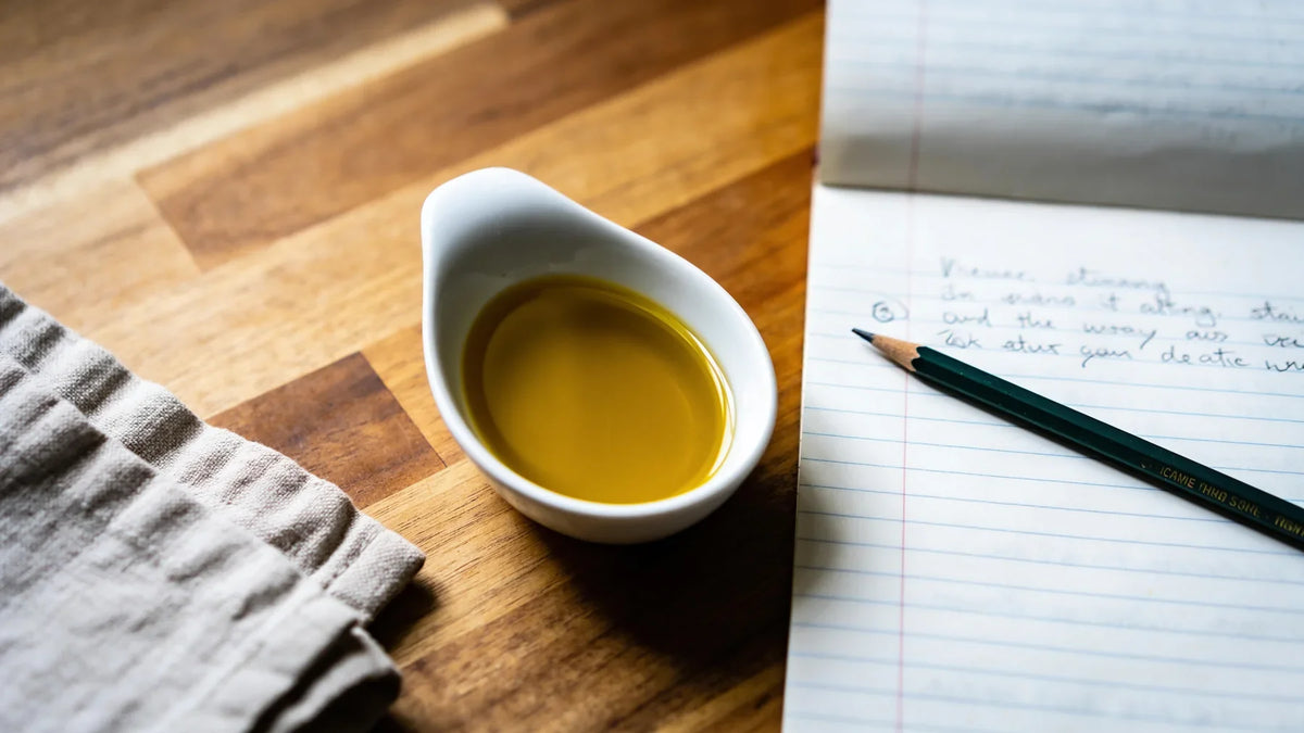 A small white dish of olive oil beside an open lined notebook with handwritten notes on a wooden table