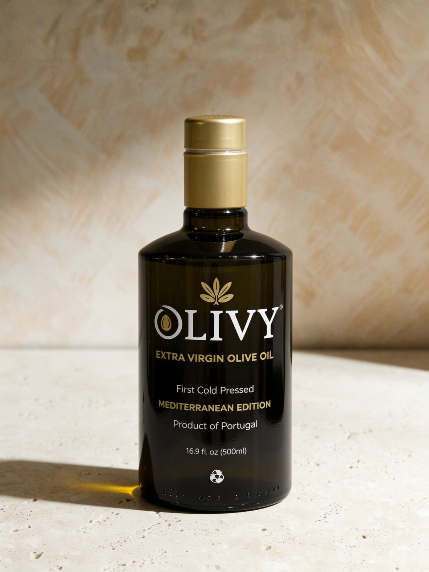 A bottle of OLIVY Extra Virgin Olive Oil, Mediterranean Edition, 16.9 fl oz (500ml), featuring a dark glass bottle with a gold cap, set against a textured beige background with dramatic side lighting.