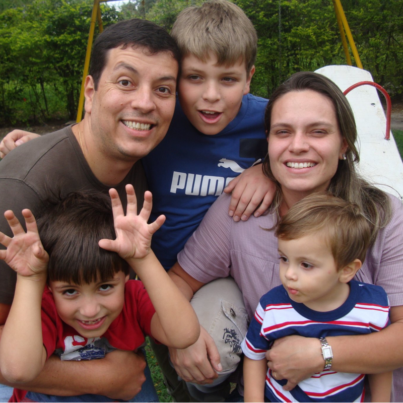 A family of five smiling outdoors on a playground with trees in the background. The father wears a brown shirt, the mother wears a light purple shirt, and the three children wear colorful Puma apparel, including a blue Puma shirt, a red shirt, and a blue and red striped shirt.