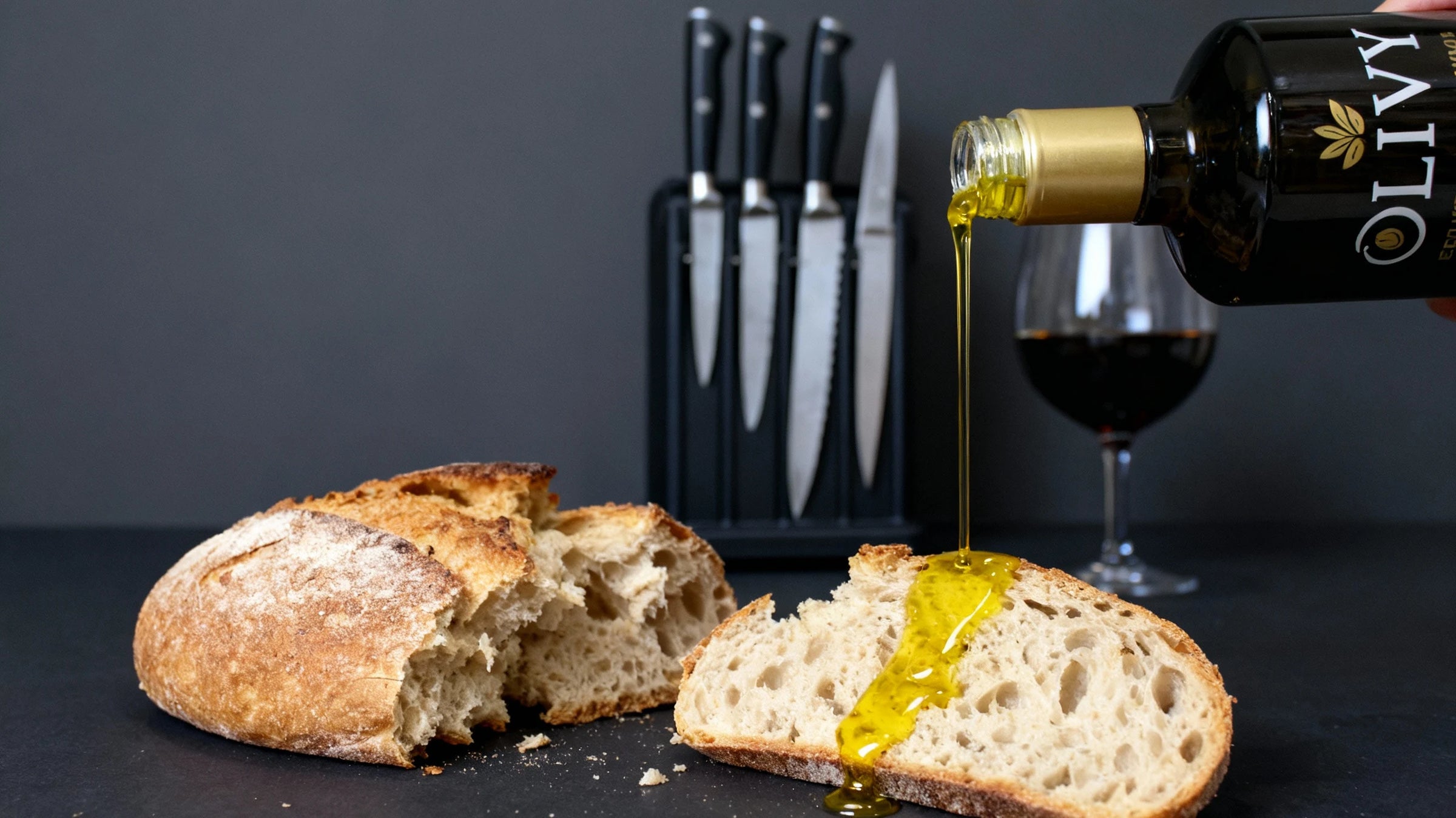 Bread with olive oil, wine glass, and knives on a dark surface