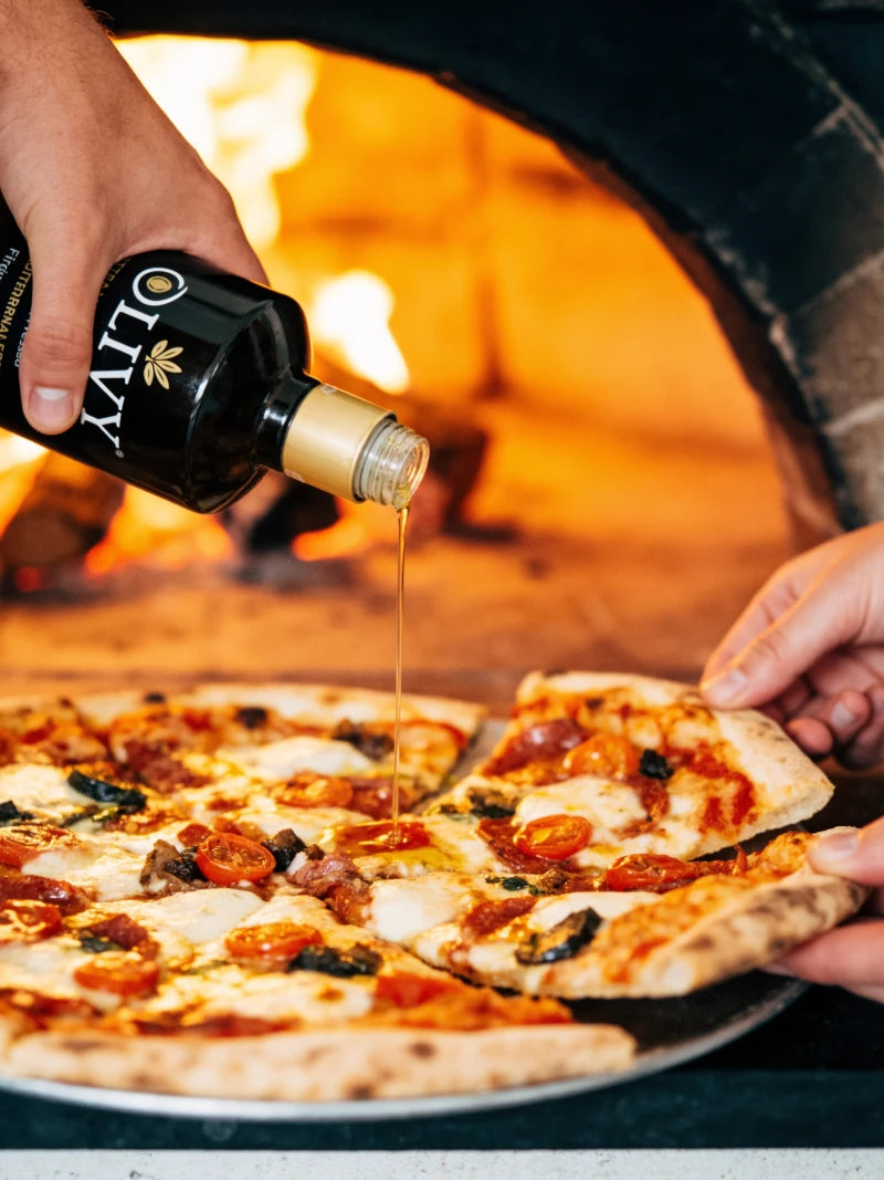 A hand pours olive oil from a black bottle onto a freshly baked pizza inside an outdoor wood-fired oven. The pizza is topped with melted cheese, sliced pepperoni, and green basil.