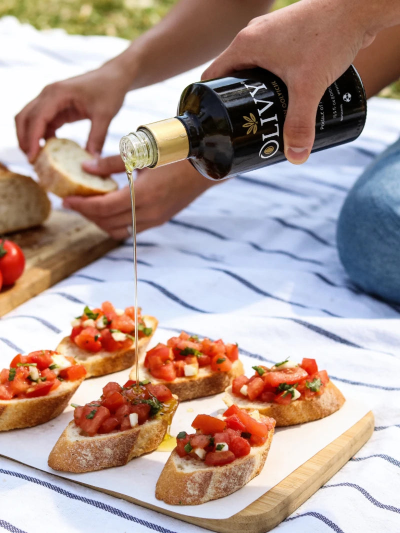 A person pouring olive oil from a black bottle with a gold collar onto bruschetta slices arranged on a wooden board over a white picnic blanket with blue stripes.