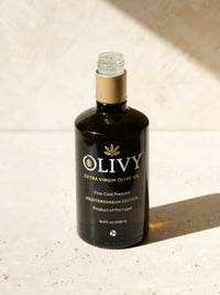 A bottle of OLIVY Extra Virgin Olive Oil, Mediterranean Edition, set on a textured beige stone surface with warm sunlight casting a soft shadow.